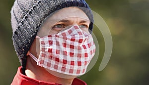 Young man in warm beanie hat wearing home made cotton face mouth nose virus mask, blurred sunny park in background, can be used