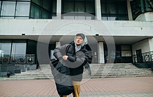 A young man in warm casual clothes stands on the street in the background of the building and pulls things out of his backpack and