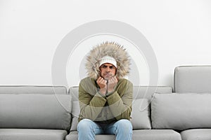 Young man in warm clothes freezing on sofa against background