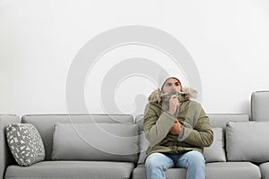 Young man in warm clothes freezing on sofa against background