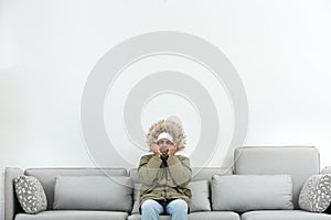 Young man in warm clothes freezing on sofa against background