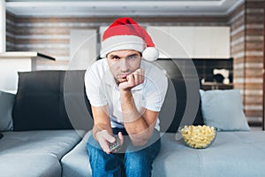 Young man watch tv in his own apartment. Bored guy with christams hat use remote control. Unhappy confused person