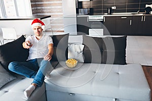 Young man watch tv in his own apartment. Picture of movieholic in christmas hat celebrate new year and watch tv and use