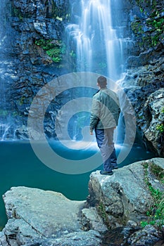 Young man watching waterfall falling from mountain with calm blurred water surface at morning