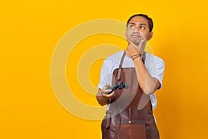 Young man wearing apron holding game controller looking aside with doubtful and skeptical expression on yellow background