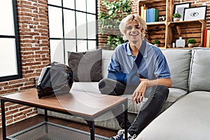 Young man wearing arm on sling for motorcycle accident looking positive and happy standing and smiling with a confident smile