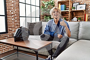 Young man wearing arm on sling for motorcycle accident smiling happy and positive, thumb up doing excellent and approval sign