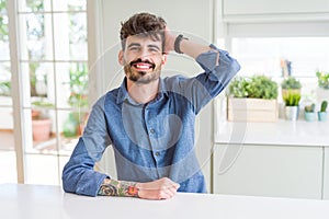 Young man wearing casual shirt sitting on white table Smiling confident touching hair with hand up gesture, posing attractive
