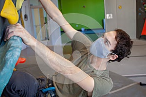 Young man wearing a COVID-19 pandemic mask climbs a bouldering wall in a climbing gym following social distance guidelines