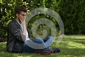 A young man wearing glasses is sitting on the grass of a park with his laptop. He is with her legs crossed and he seems to be