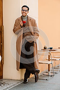 Young man wearing glasses smiling and drinking coffee on city street