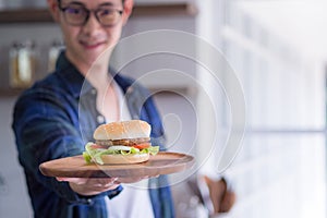 Young man wearing glasses smiling, looking at the camera, and holding a homemade hamburger on a wooden dish with blur background.