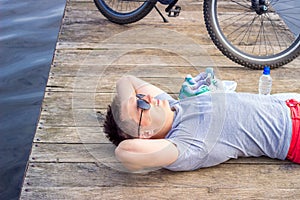 Young man wearing in gray shirt with glasses listening music headphones lying at the beach and resting after bicycle ride