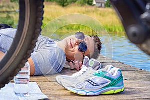 Young man wearing in gray shirt with glasses listening music headphones lying at the beach and resting after bicycle ride