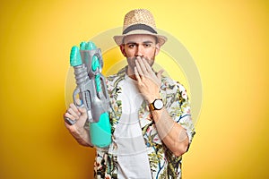 Young man wearing hawaiian flowers shirt holding water gun over yellow isolated background cover mouth with hand shocked with