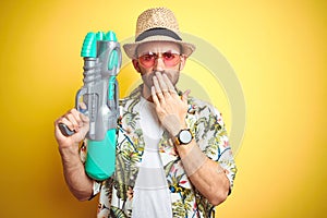 Young man wearing hawaiian flowers shirt holding water gun over yellow isolated background cover mouth with hand shocked with