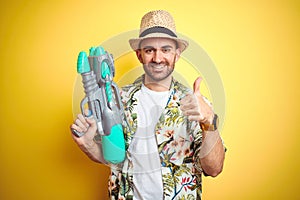 Young man wearing hawaiian flowers shirt holding water gun over yellow isolated background happy with big smile doing ok sign,