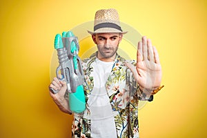 Young man wearing hawaiian flowers shirt holding water gun over yellow isolated background with open hand doing stop sign with