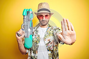 Young man wearing hawaiian flowers shirt holding water gun over yellow isolated background with open hand doing stop sign with