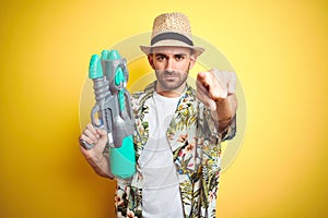 Young man wearing hawaiian flowers shirt holding water gun over yellow isolated background pointing with finger to the camera and
