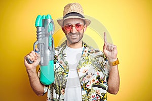 Young man wearing hawaiian flowers shirt holding water gun over yellow isolated background surprised with an idea or question