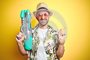 Young man wearing hawaiian flowers shirt holding water gun over yellow isolated background very happy pointing with hand and