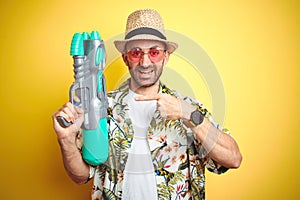 Young man wearing hawaiian flowers shirt holding water gun over yellow isolated background very happy pointing with hand and
