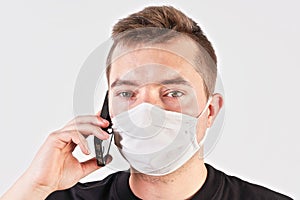 Young man wearing home made cotton face mouth nose virus mask talking on his mobile phone. Studio portrait, white background  -