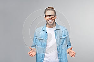 Young man wearing jeans shirt welcoming you with a smile on his face and his arms wide open standing over grey