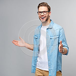 Young man wearing jeans shirt welcoming you with a smile on his face and his arms wide open standing over grey