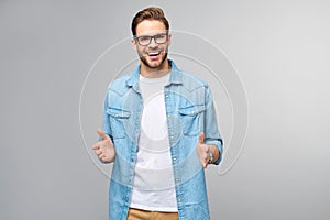 Young man wearing jeans shirt welcoming you with a smile on his face and his arms wide open standing over grey