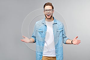 Young man wearing jeans shirt welcoming you with a smile on his face and his arms wide open standing over grey