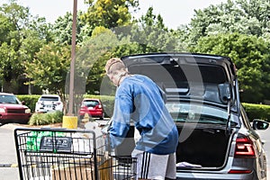 Young man wearing mask loads groceries from cart to trunk of car in parking lot during pandemic