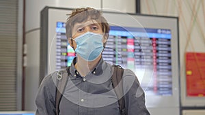 A young man wearing a medical face mask in an airport stands in a hall in front of a flight schedule monitor. The