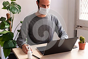 Young man wearing medical face mask at work from home due to corona virus outbreak with laptop and mobile phone on wooden table, photo