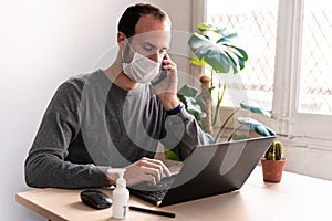 Young man wearing medical face mask at work from home due to corona virus outbreak with laptop and mobile phone on wooden table, photo