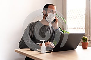 Young man wearing medical face mask at work from home due to corona virus outbreak with laptop and mobile phone on wooden table, photo