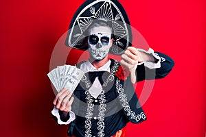 Young man wearing mexican day of the dead costume holding dollars with angry face, negative sign showing dislike with thumbs down,