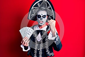 Young man wearing mexican day of the dead costume holding dollars annoyed and frustrated shouting with anger, yelling crazy with
