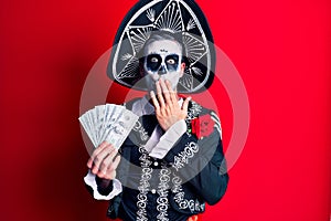 Young man wearing mexican day of the dead costume holding dollars covering mouth with hand, shocked and afraid for mistake