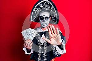 Young man wearing mexican day of the dead costume holding dollars with open hand doing stop sign with serious and confident