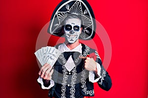 Young man wearing mexican day of the dead costume holding dollars pointing finger to one self smiling happy and proud