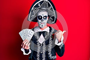 Young man wearing mexican day of the dead costume holding dollars pointing thumb up to the side smiling happy with open mouth