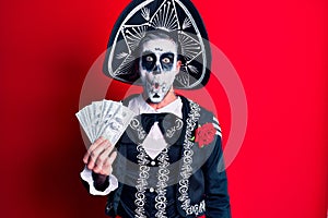 Young man wearing mexican day of the dead costume holding dollars scared and amazed with open mouth for surprise, disbelief face