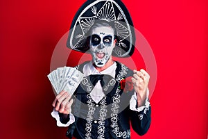 Young man wearing mexican day of the dead costume holding dollars screaming proud, celebrating victory and success very excited