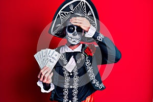Young man wearing mexican day of the dead costume holding dollars stressed and frustrated with hand on head, surprised and angry