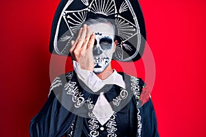 Young man wearing mexican day of the dead costume over red covering one eye with hand, confident smile on face and surprise