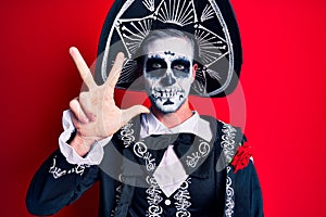 Young man wearing mexican day of the dead costume over red showing and pointing up with fingers number three while smiling