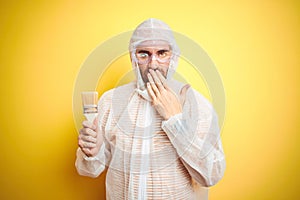 Young man wearing painter equipment and holding painting brush over isolated yellow background cover mouth with hand shocked with