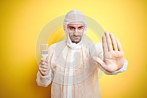 Young man wearing painter equipment and holding painting brush over isolated yellow background with open hand doing stop sign with